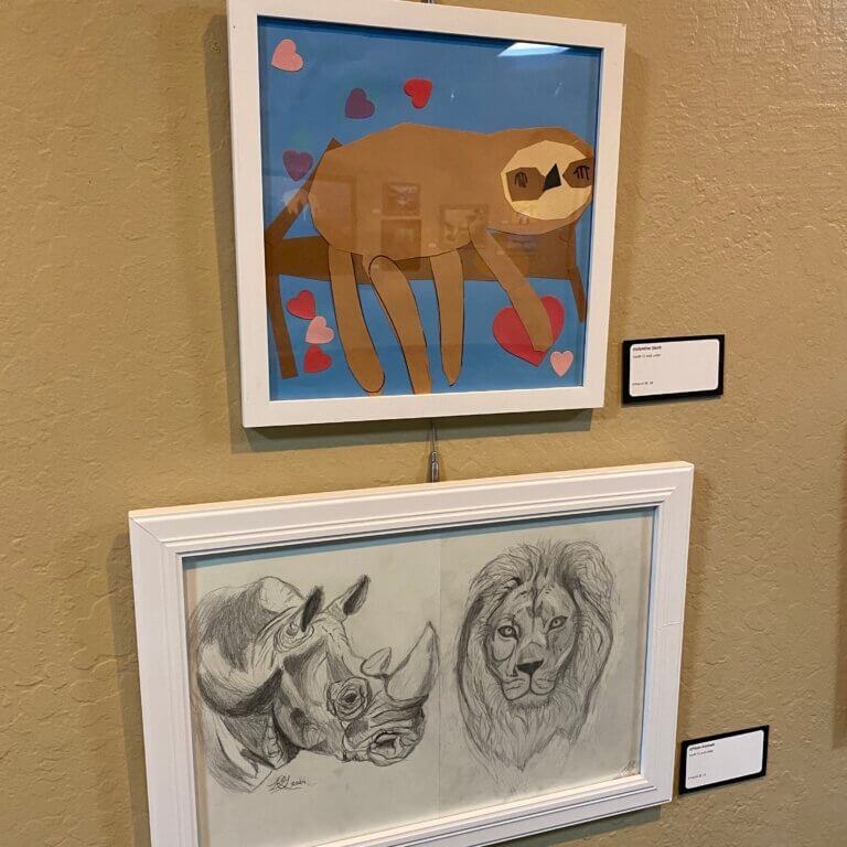 Photo of two pieces of framed youth artwork. One is a paper craft of a sloth, and the other is a drawing of a lion and a rhinoceros.