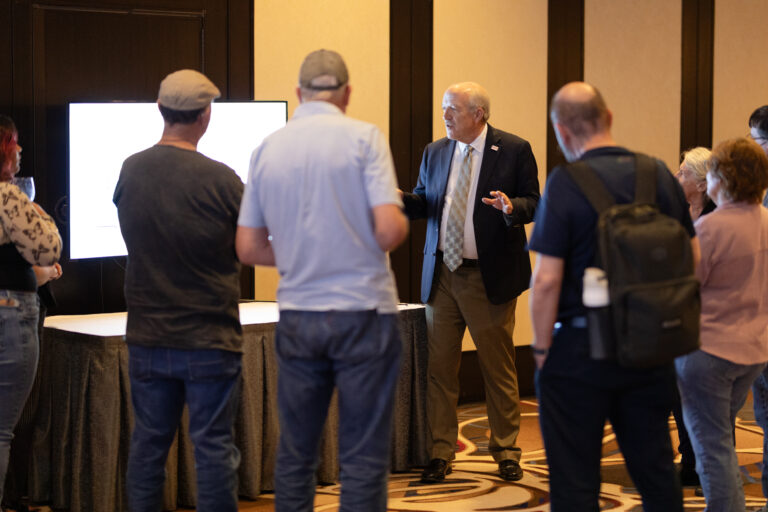 Photo of a speaker presentation in front of a screen with people standing around